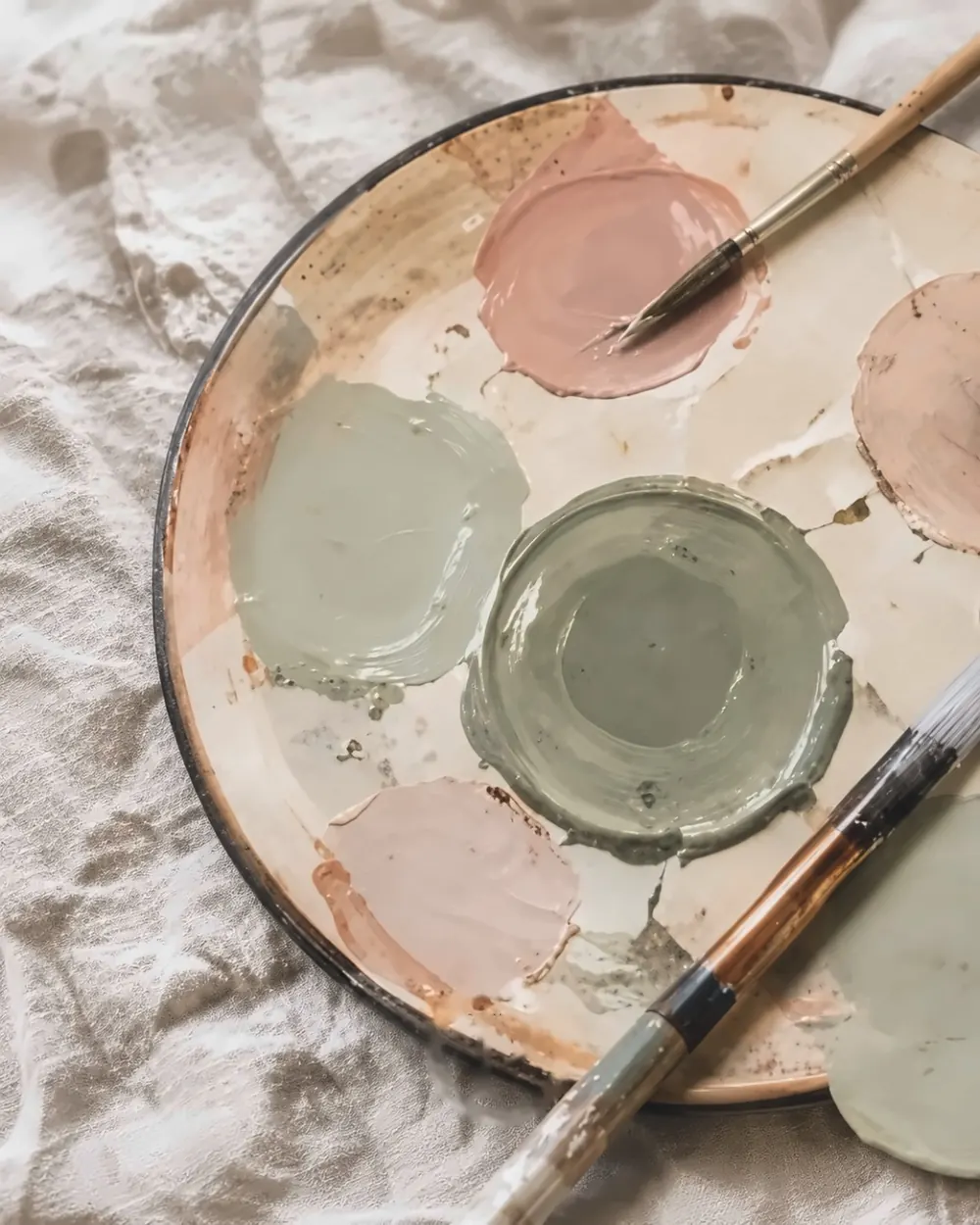 Farbpalette mit Salbei- und Rosétönen und zwei Pinseln auf hellem Leinen
